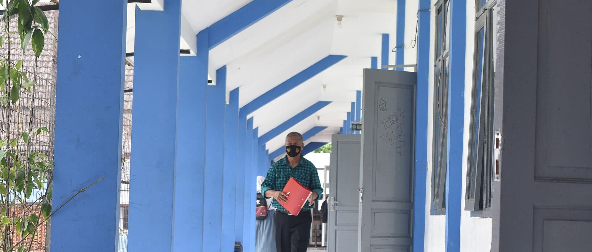 Seorang guru berjalan menyusuri koridor sekolah bertiang biru sambil membawa buku